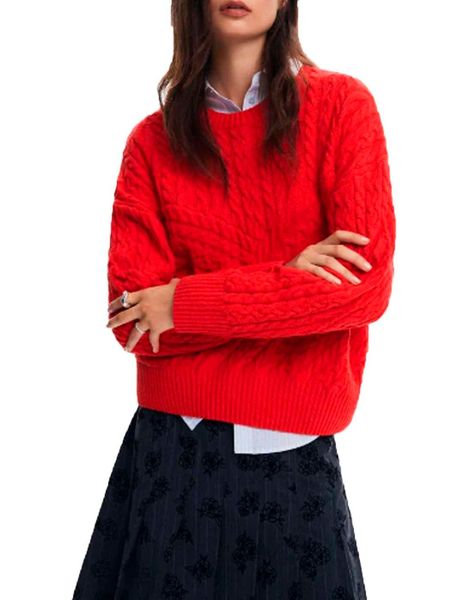 Donna con maglione rosso a trecce, braccia incrociate, sopra una gonna blu a fiori.