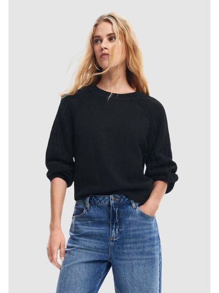 Donna che indossa un maglione nero e jeans blu, in piedi in casa con le mani in tasca.