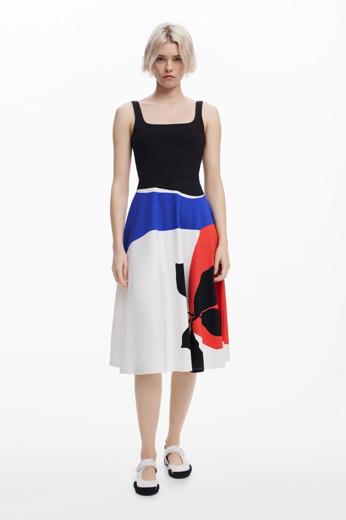 Donna con top nero e gonna colorata, scarpe bianche, in piedi davanti a uno sfondo bianco.