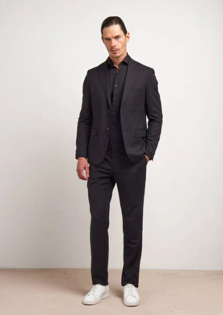 Un uomo in abito nero, camicia nera e scarpe da ginnastica bianche è in piedi su uno sfondo semplice.