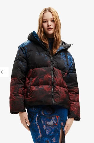 Donna con piumino fantasia con cappuccio sopra un abito blu. Sfumature di nero, rosso e blu.