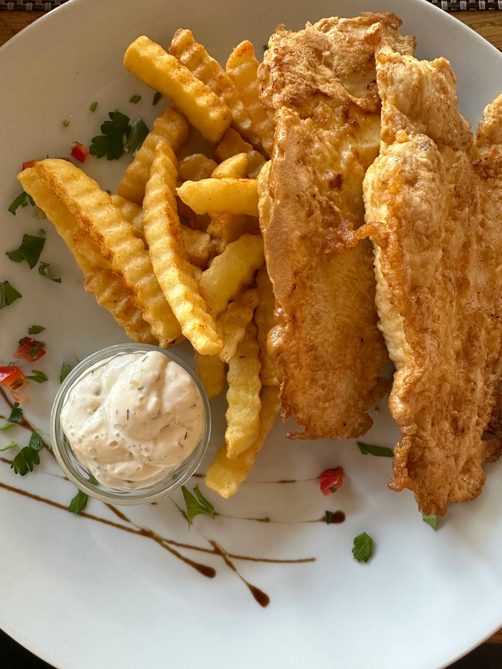 Fish and chips with tartar sauce on a white plate, garnished with herbs and a drizzle.