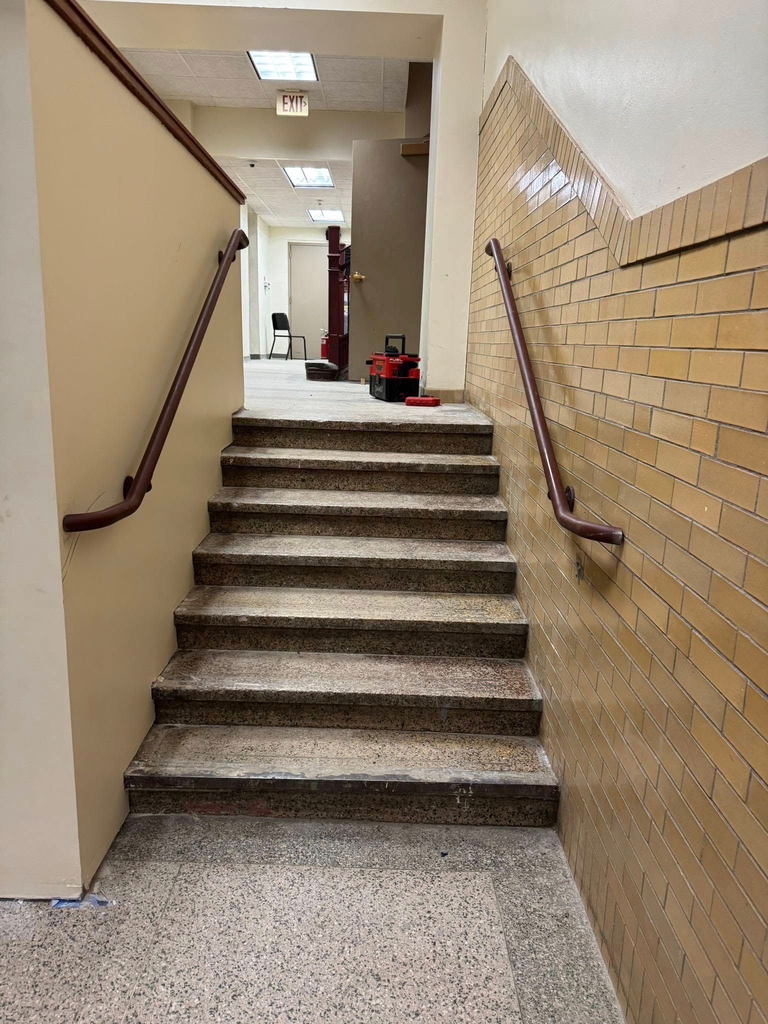 Staircase with brown handrails and light-colored walls. A red utility cart sits at the top of the stairs.