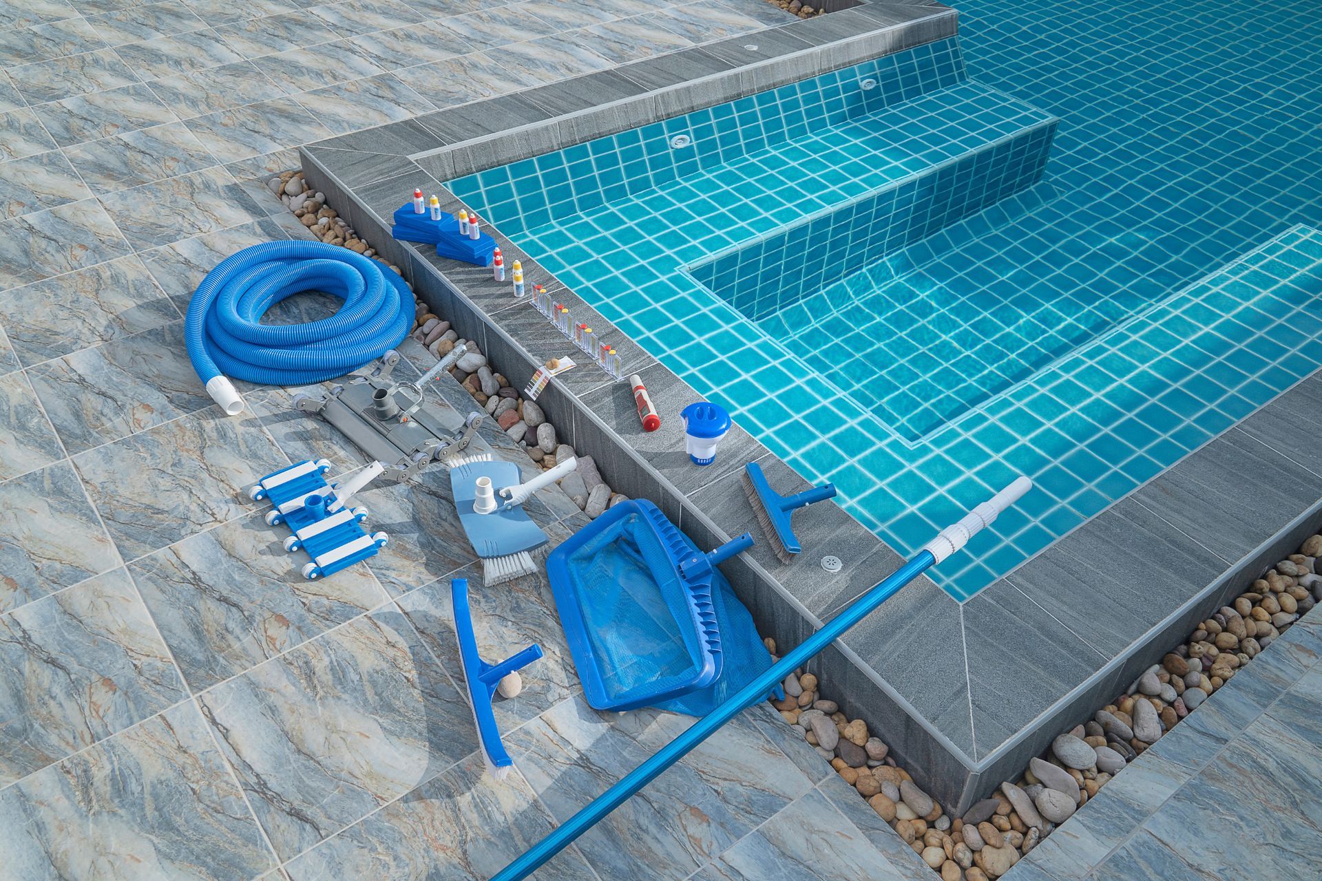 Pool and cleaning supplies, including a vacuum hose, skimmer, brushes, and net, sit on a pool deck.