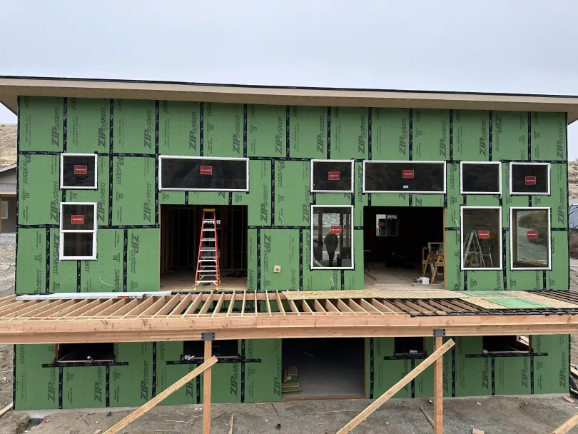 Green modular house under construction with exposed framing, windows, and a wooden deck scaffolding.