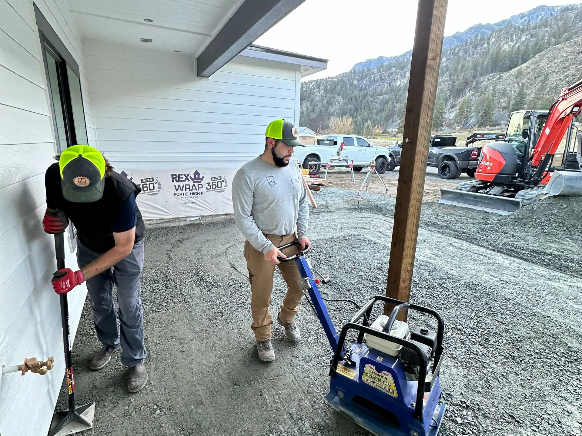 Two workers with shovels and a plate compactor clearing gravel beside a white building