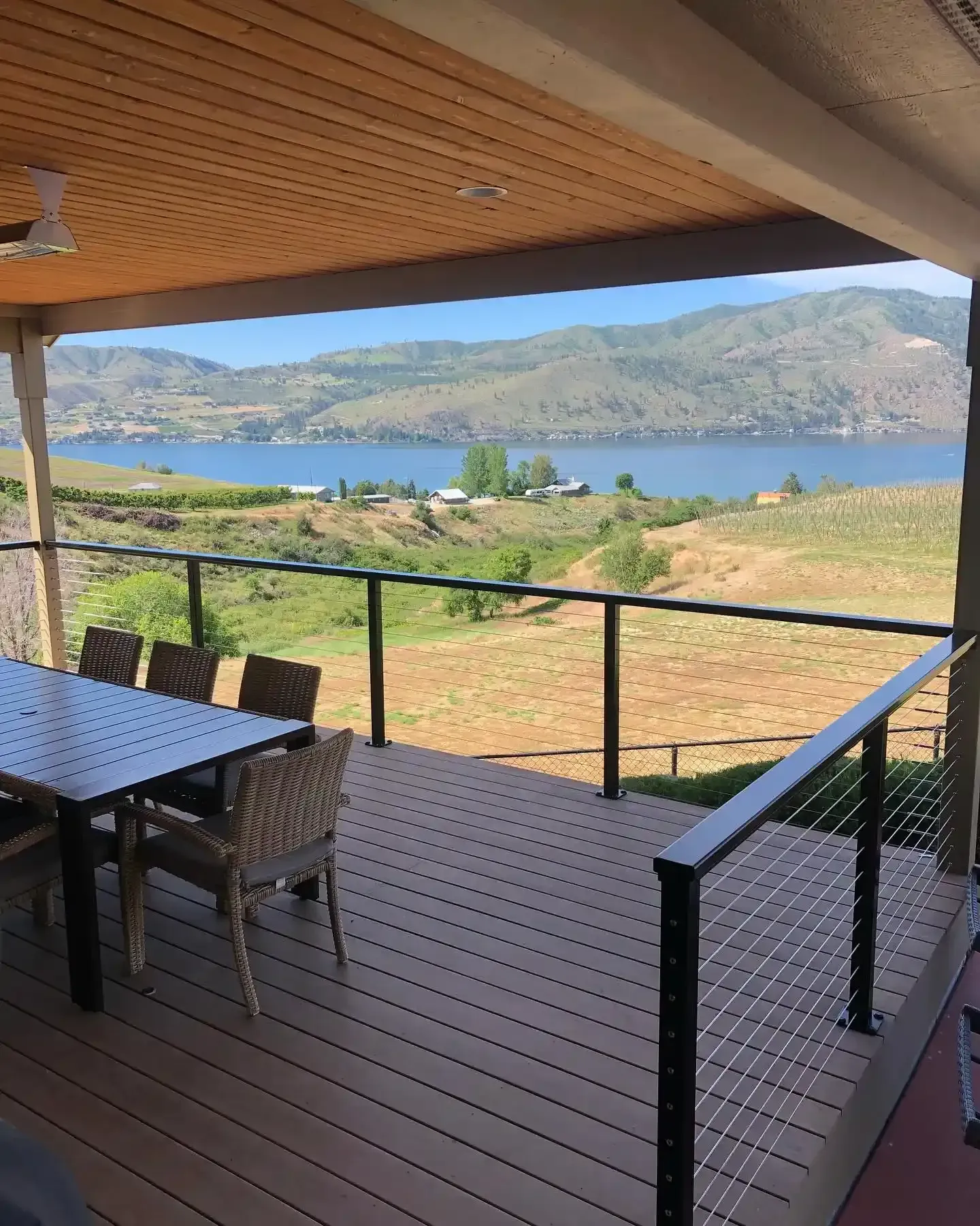 Covered deck overlooking a lake and green hills, with patio table, chairs, and railing in the foreground