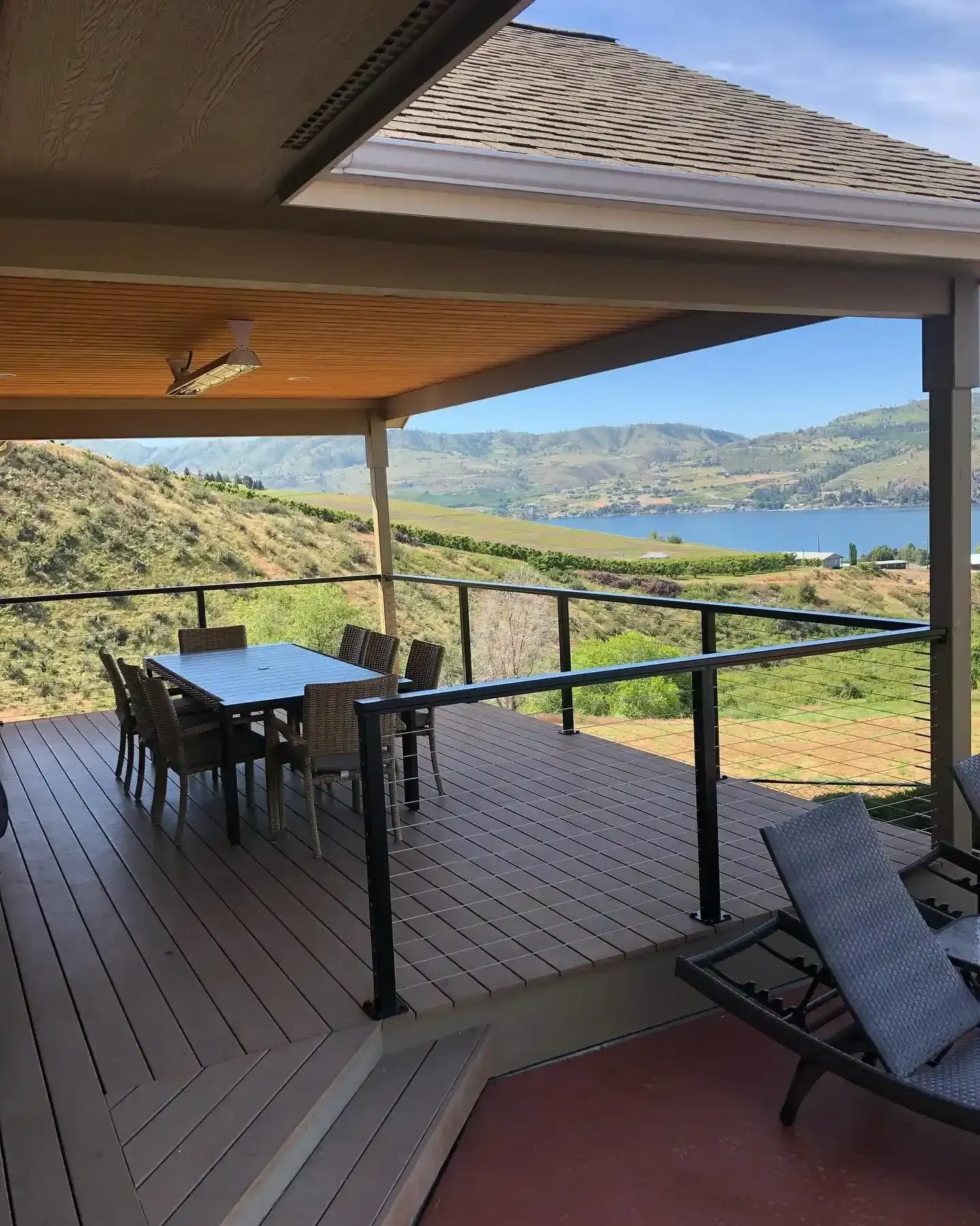 Covered deck with table and chairs overlooking a lake and green hills.