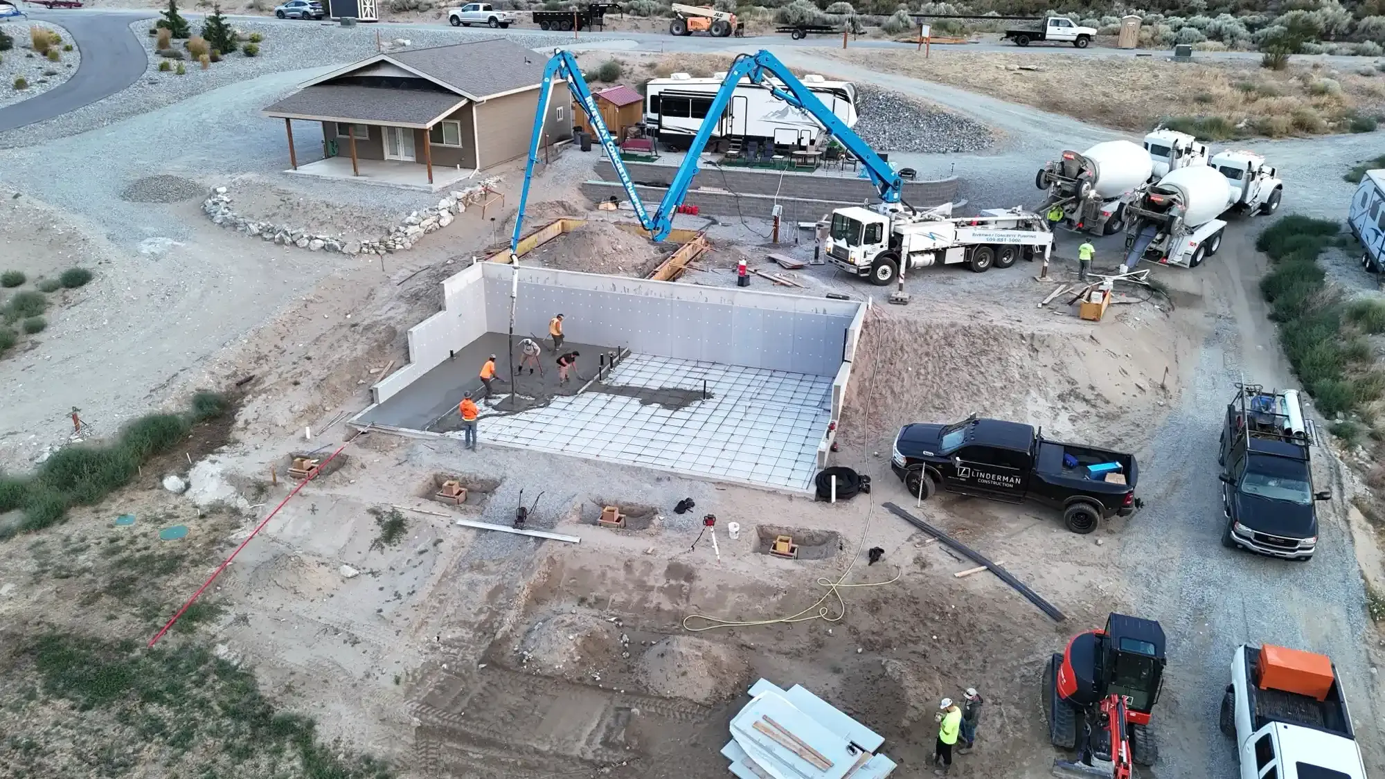 Aerial view of a construction site with a concrete foundation, blue excavators, and several trucks.