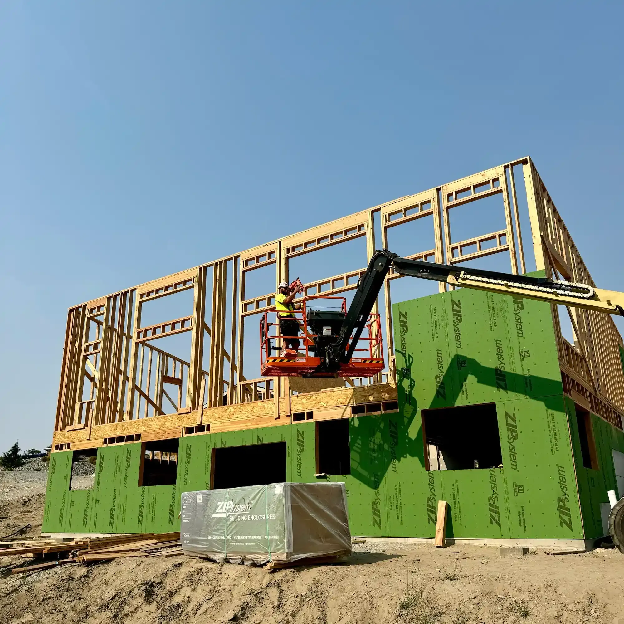 Construction site with green-wrapped house framing and a worker on a lift under a clear blue sky