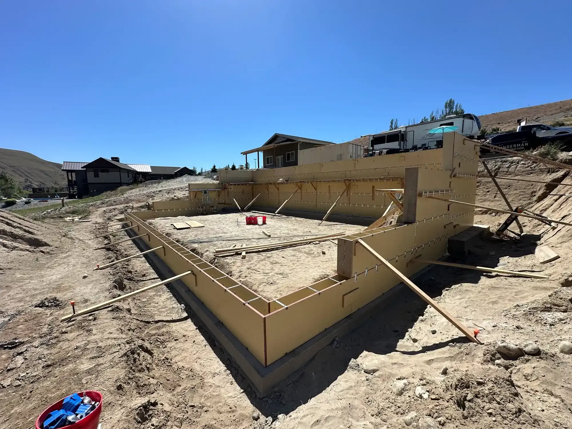 Damaged house foundation in a dirt lot under construction, with debris and a red bucket under a blue sky