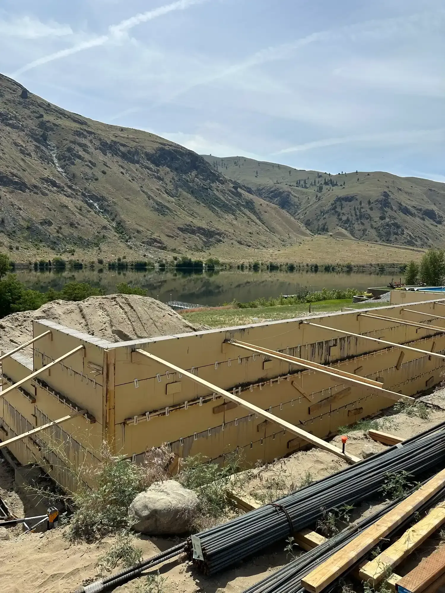 Construction site with wooden foundation forms and pipes beside a river, with mountains in the background