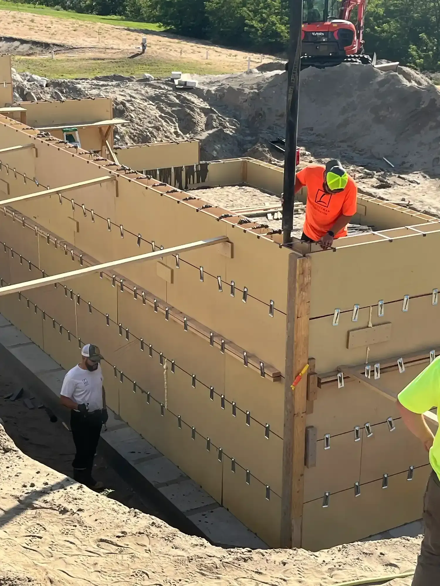 Workers building a large tan modular structure at a construction site, with dirt and a red excavator nearby