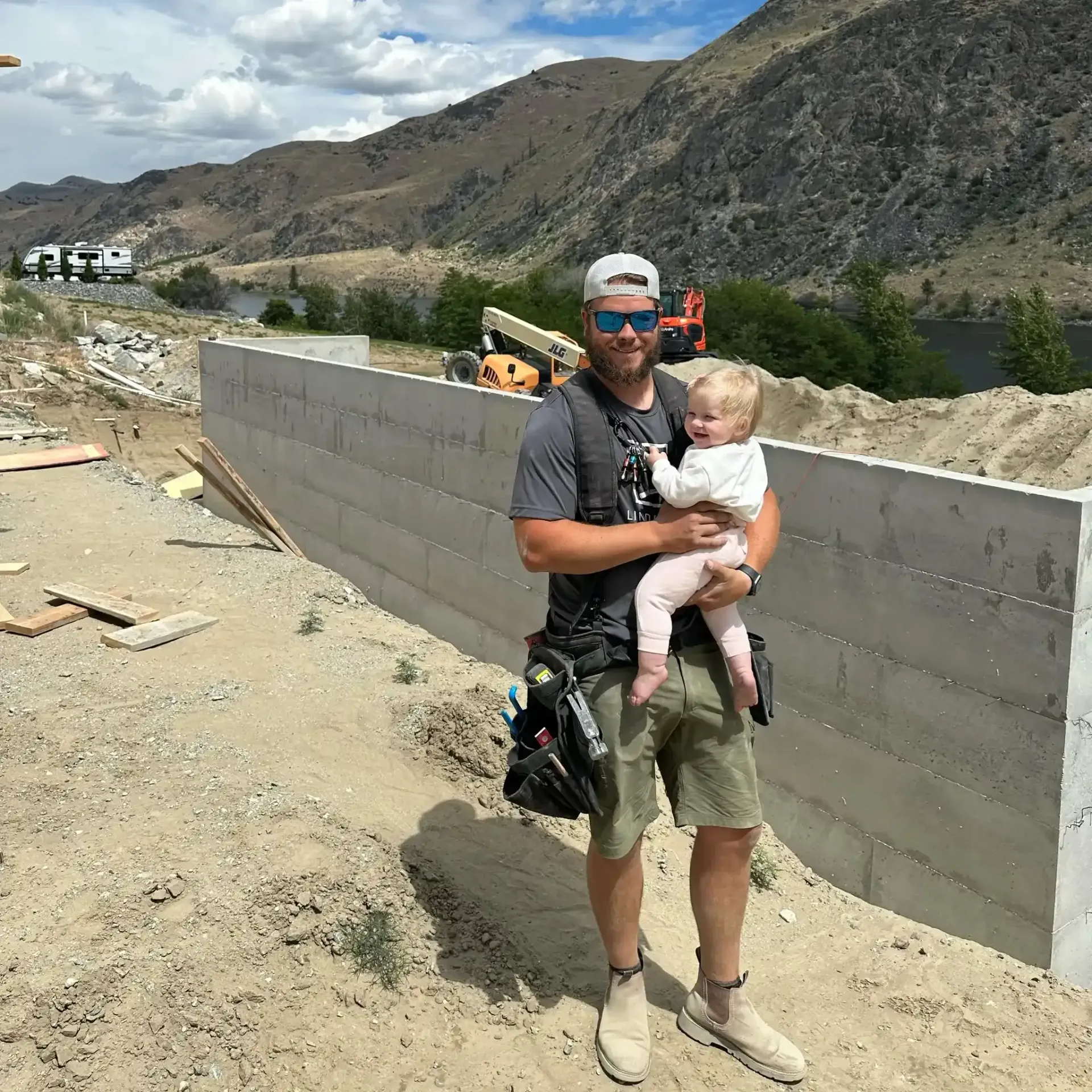 Man holding a baby beside a cinder block wall at a rocky construction site with mountains in the background