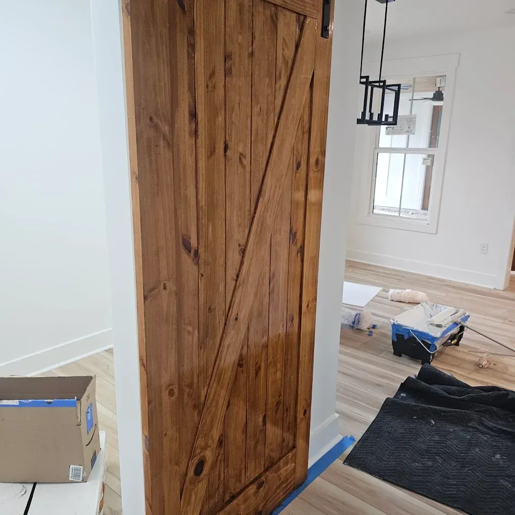 Wooden barn door, stained brown, installed in a white-walled doorway. Light streams from a window nearby.