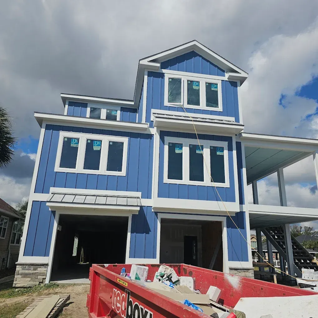 Blue house under construction with white trim, a red dumpster, and a partially cloudy sky.
