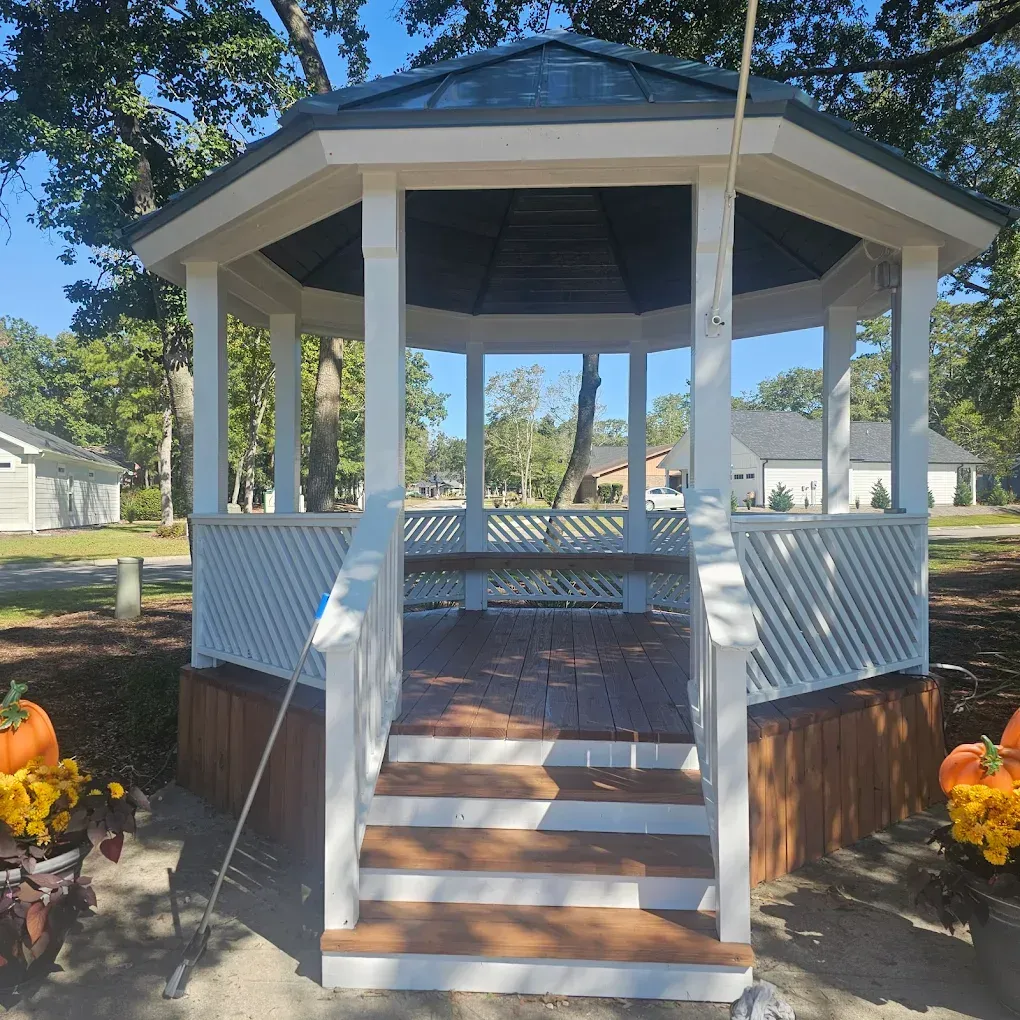 White gazebo with a dark roof and wooden deck. Surrounded by a lattice railing and steps.