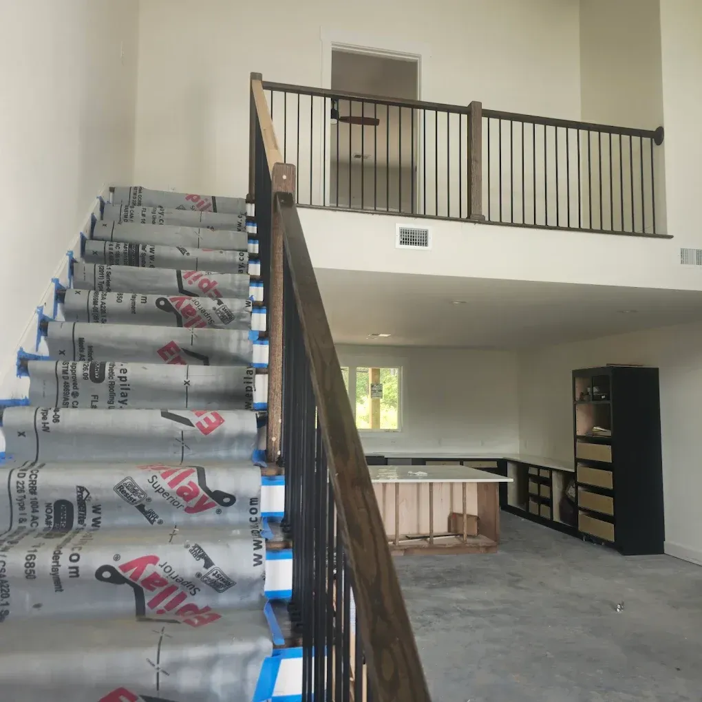 Interior view of a two-story home under construction. A staircase with protective covering leads up to a balcony.