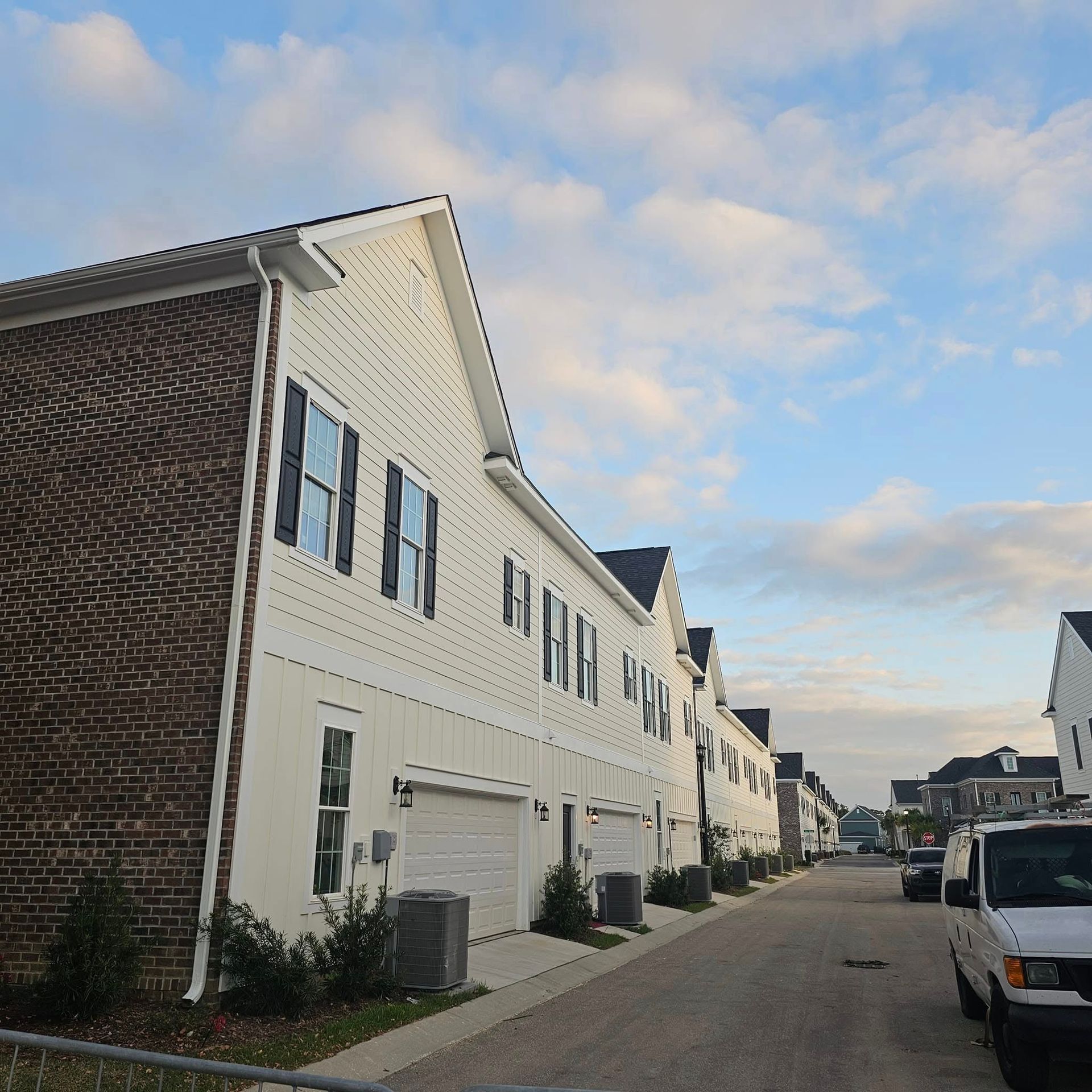 Row of white townhouses with black shutters and brick accents on a street with a cloudy sky.