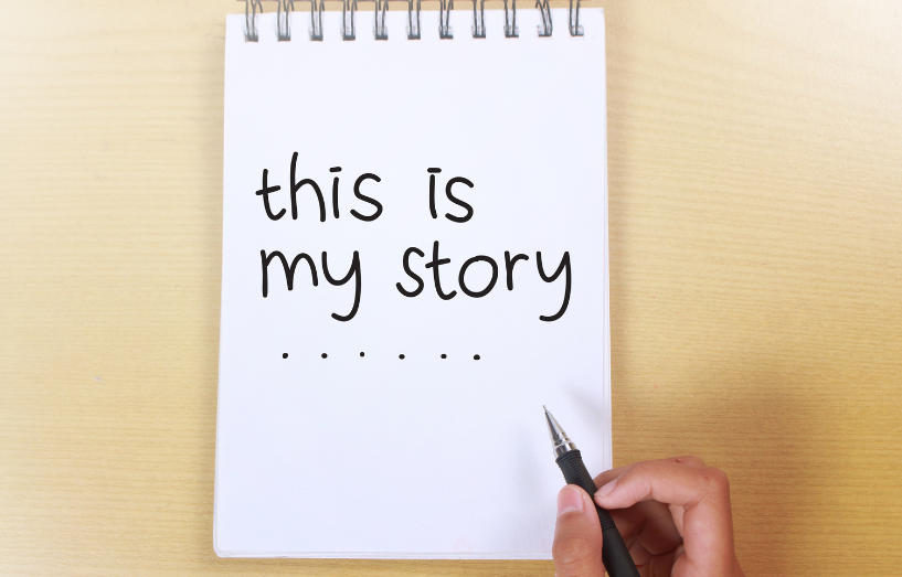 Notebook page reading “this is my story” with dotted line, as a hand holds a pen ready to write
