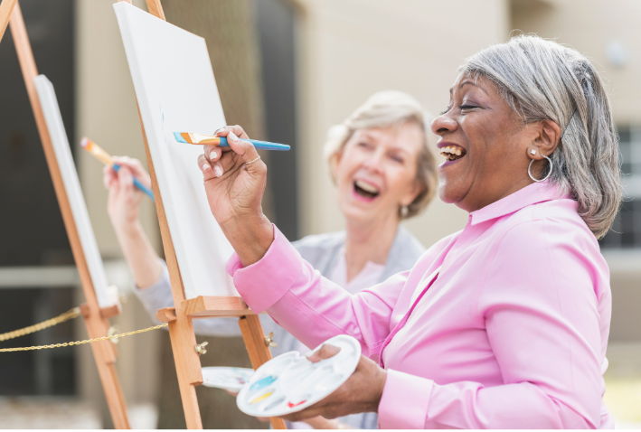 Two women painting at easels, smiling in a bright art studio.
