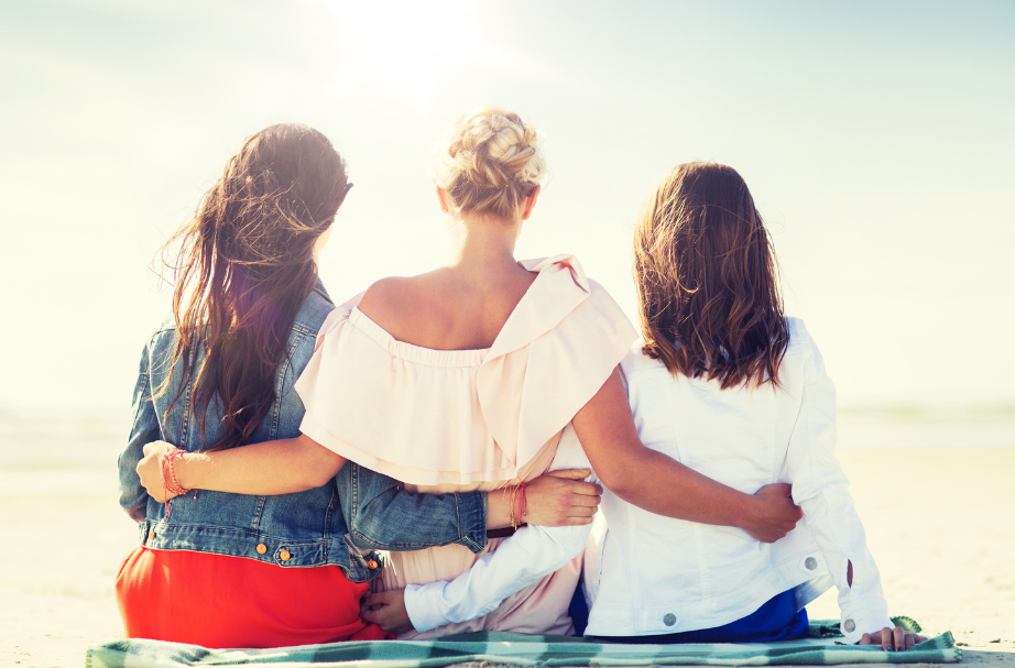 Three friends sitting on a bench with arms around each other, facing a bright beach sunset