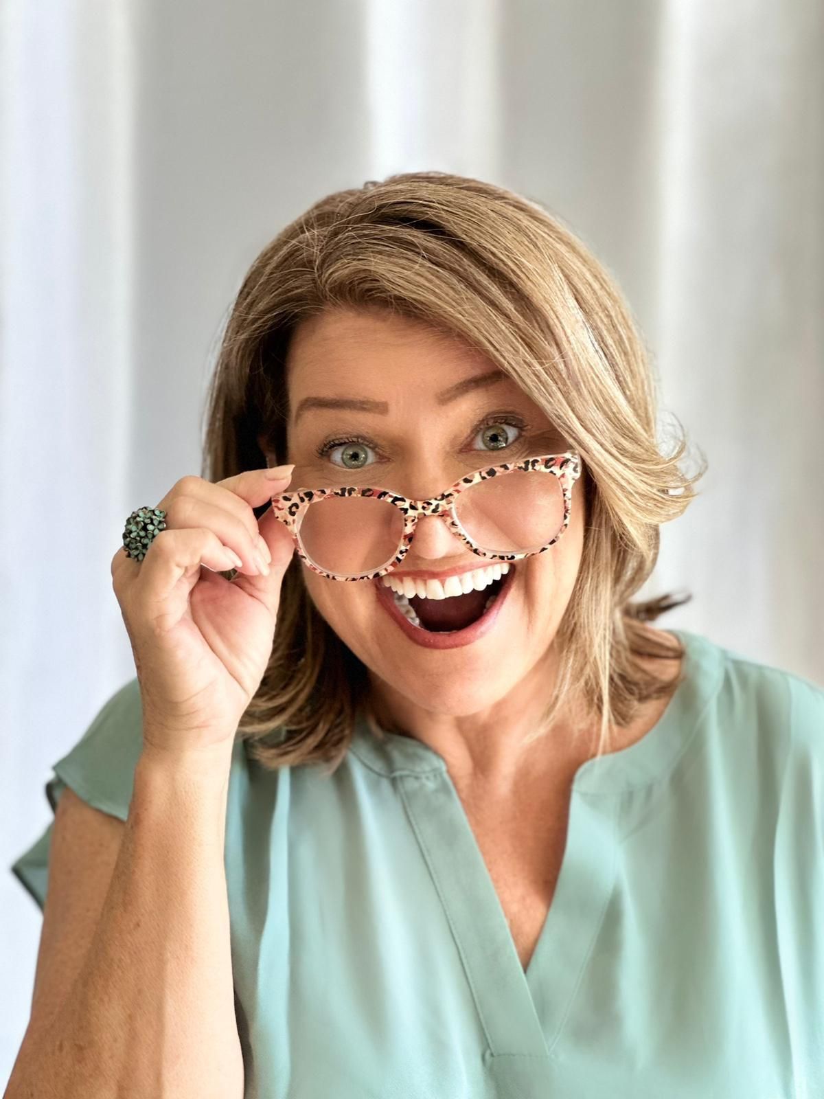 Woman in a light blue blouse lifting glasses and smiling in front of white curtains