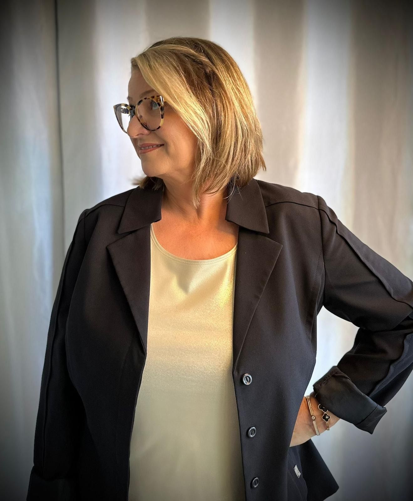 Woman in glasses wearing a dark blazer over a light top, posing against a softly lit curtain backdrop.