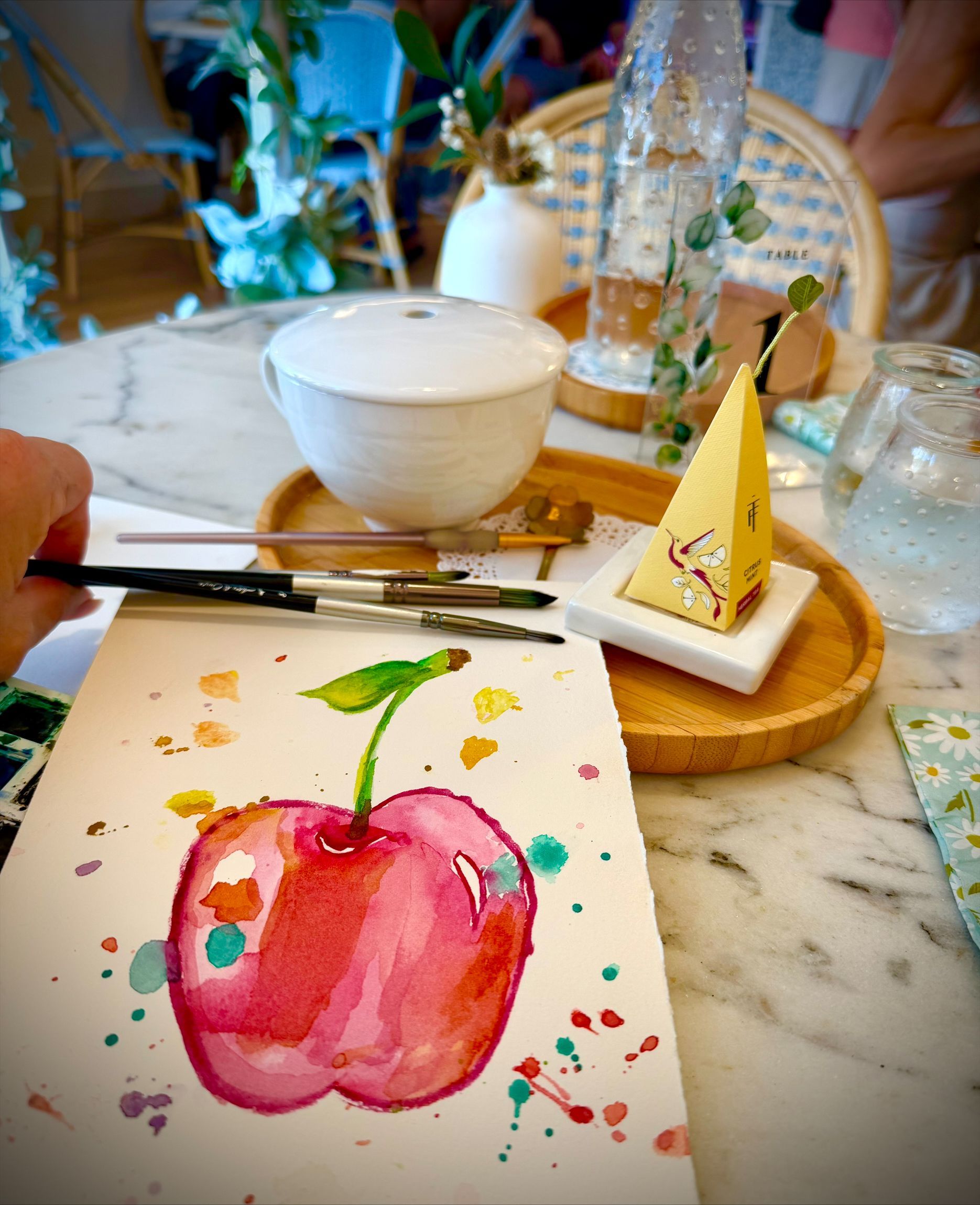 Watercolor apple painting on a table with brushes, tea set, and a dessert in a cozy café setting