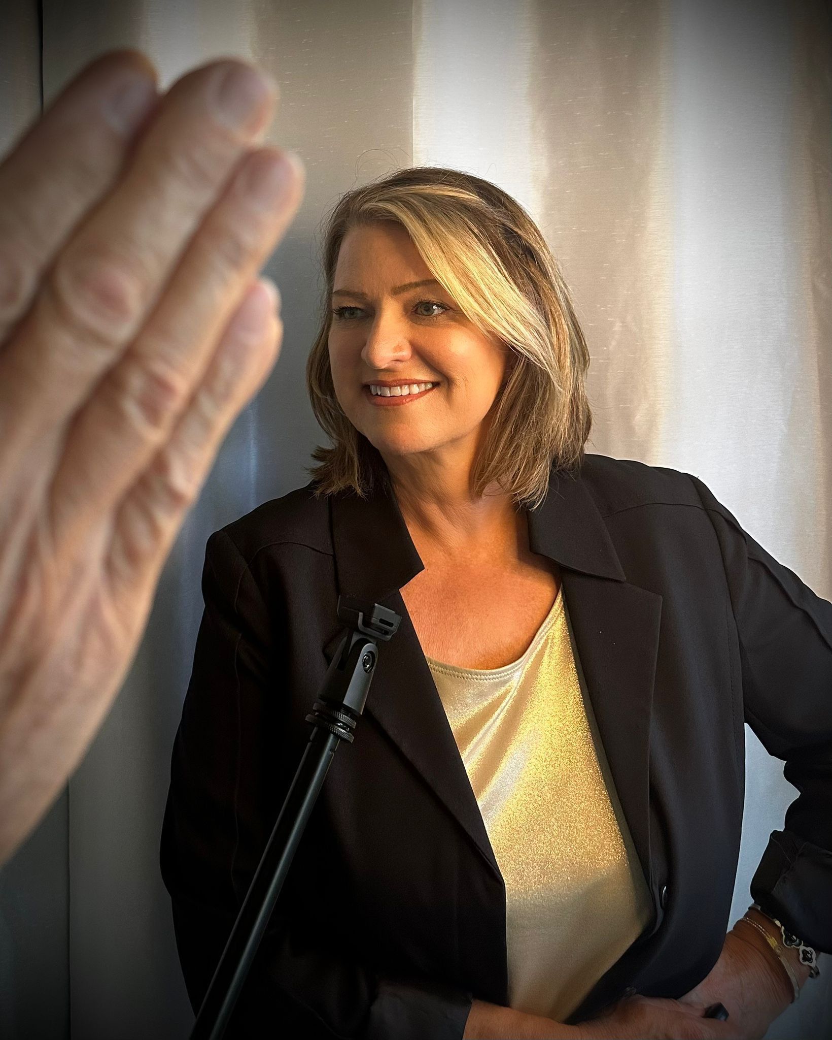 Smiling woman in a black blazer and gold top speaking at a microphone, with a raised hand in the foreground