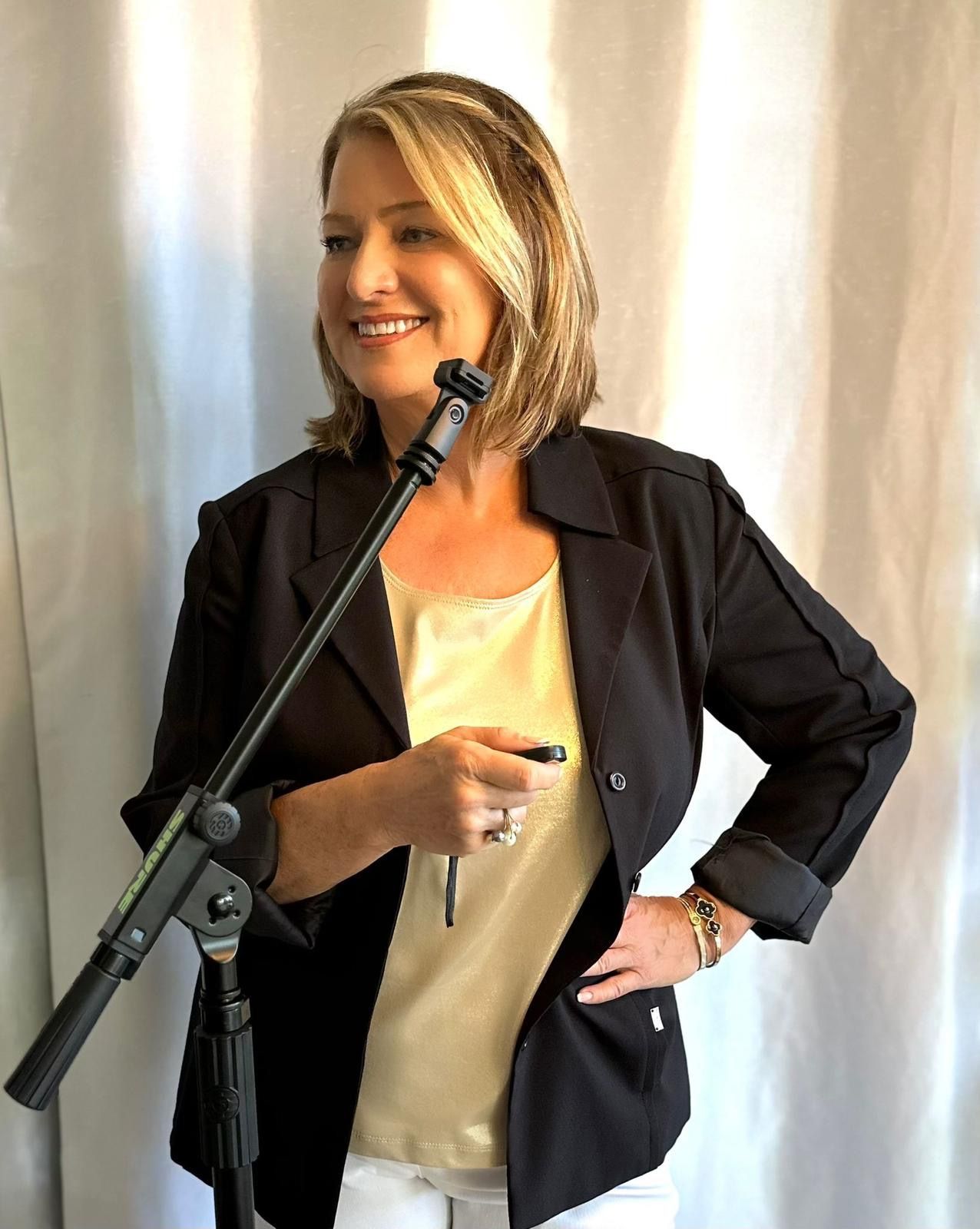 Woman smiling at a microphone, wearing a black blazer and cream top against a light curtain backdrop