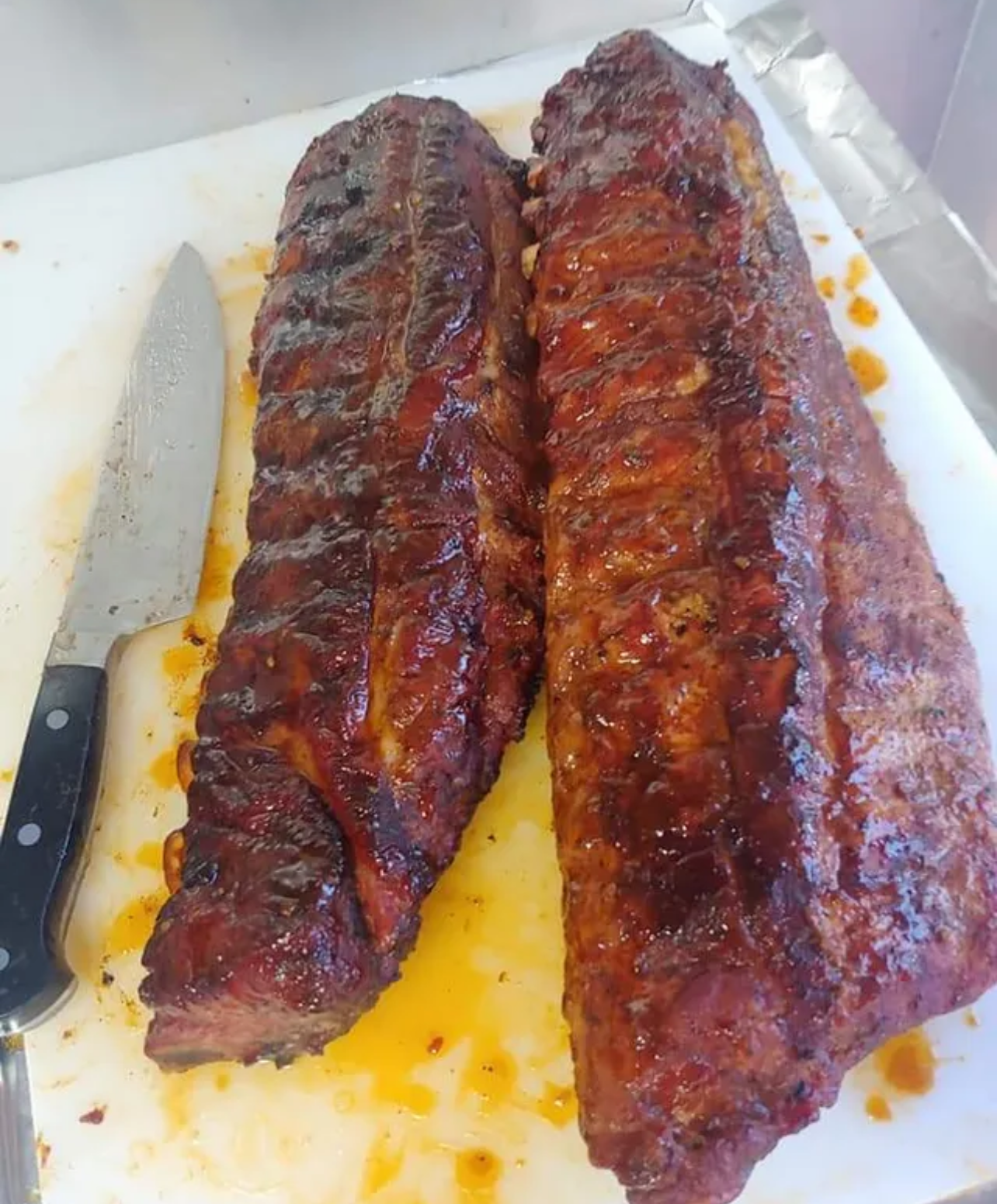 Two racks of barbecued ribs on a white cutting board next to a knife.