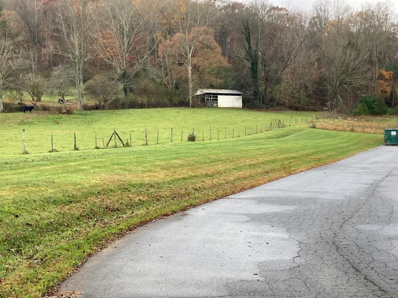 Curving wet road beside a green field and wooded hillside, with a small white shed in the distance
