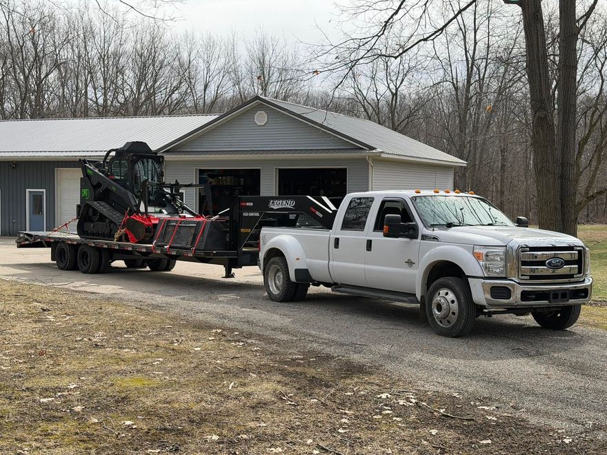 Silver pickup truck towing equipment trailer in front of a house and garage, wooded yard behind.