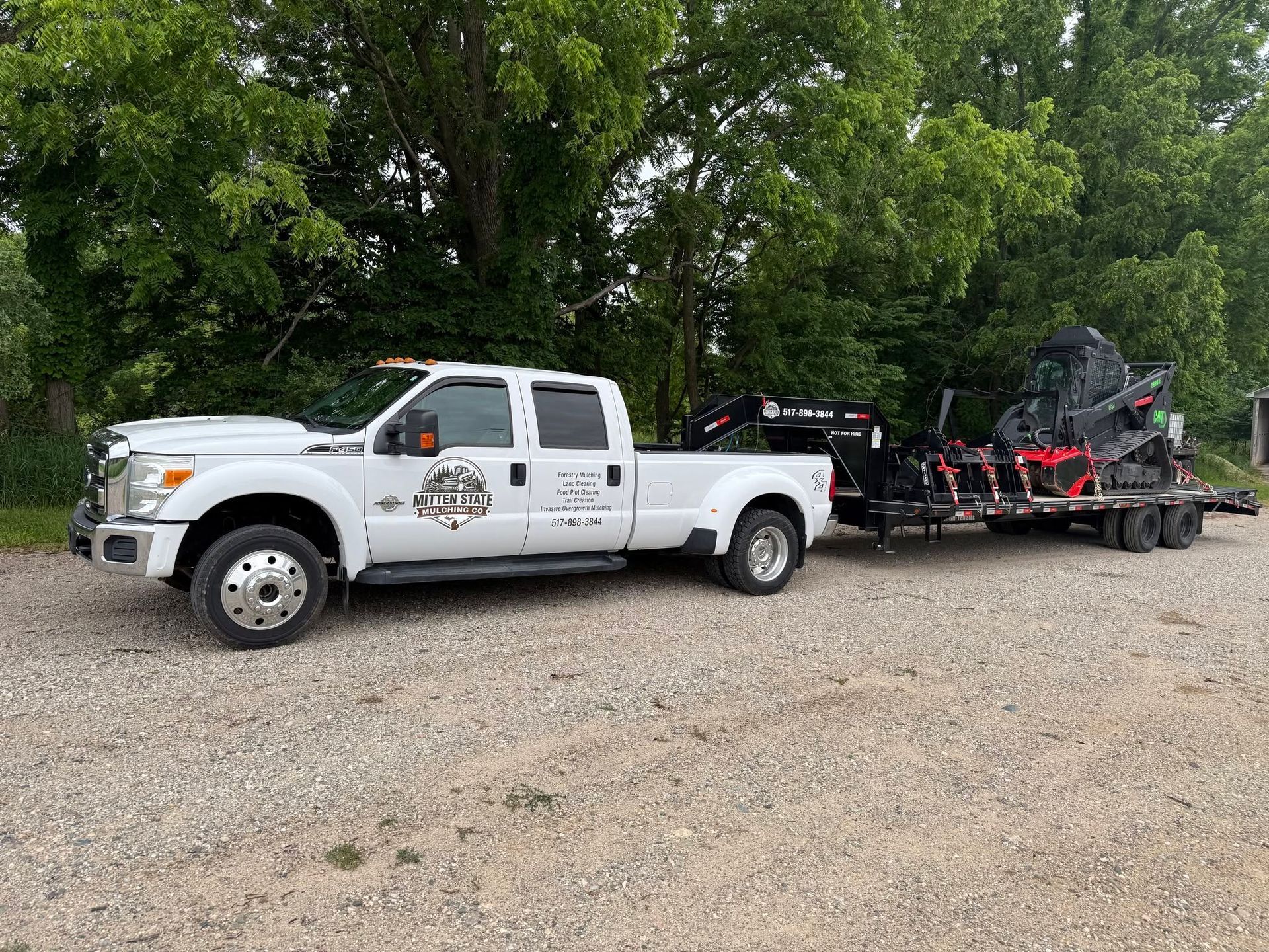White work pickup truck towing a trailer with equipment on a gravel lot beside trees