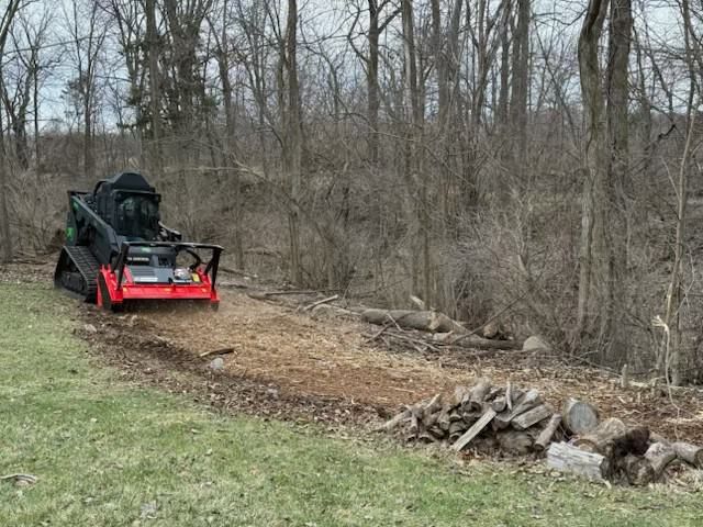 Red skid steer grading a dirt path beside a grassy area and leafless woods.