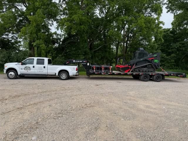 White pickup truck towing a flatbed trailer with a red-and-black skid steer on a gravel lot