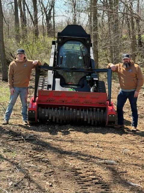 Two people standing beside a skid steer with a red mulcher in a wooded area