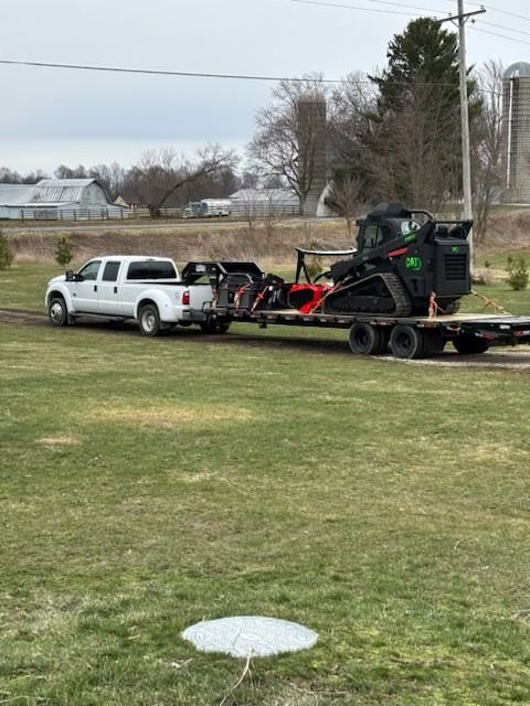 White pickup towing red trailer with black skid-steer on a grassy field near a road