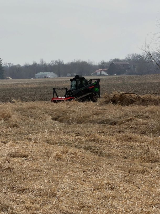 Rider on a green tractor mowing a dry field near a rural treeline
