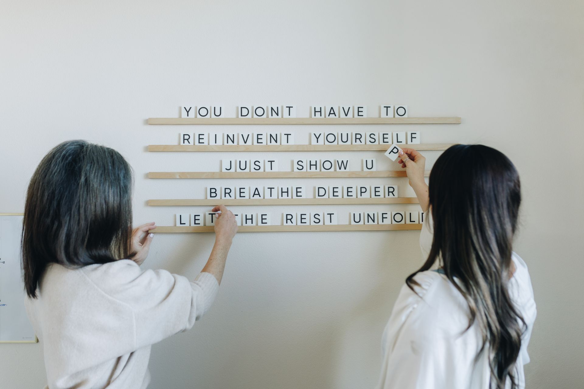 Two people place letter strips on a white wall, forming a motivational quote.