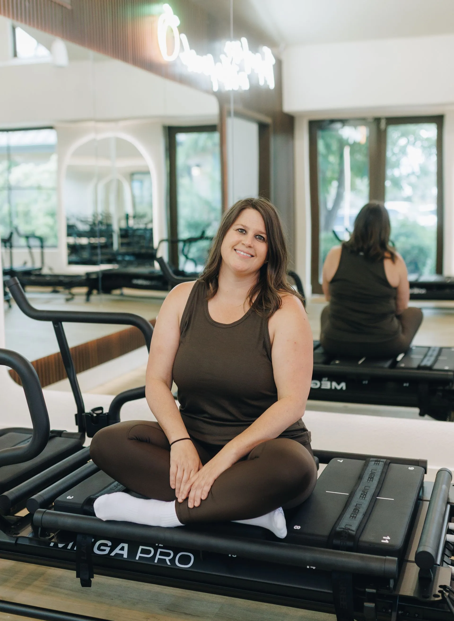 Woman sitting cross-legged on a treadmill in a bright gym, smiling at the camera.