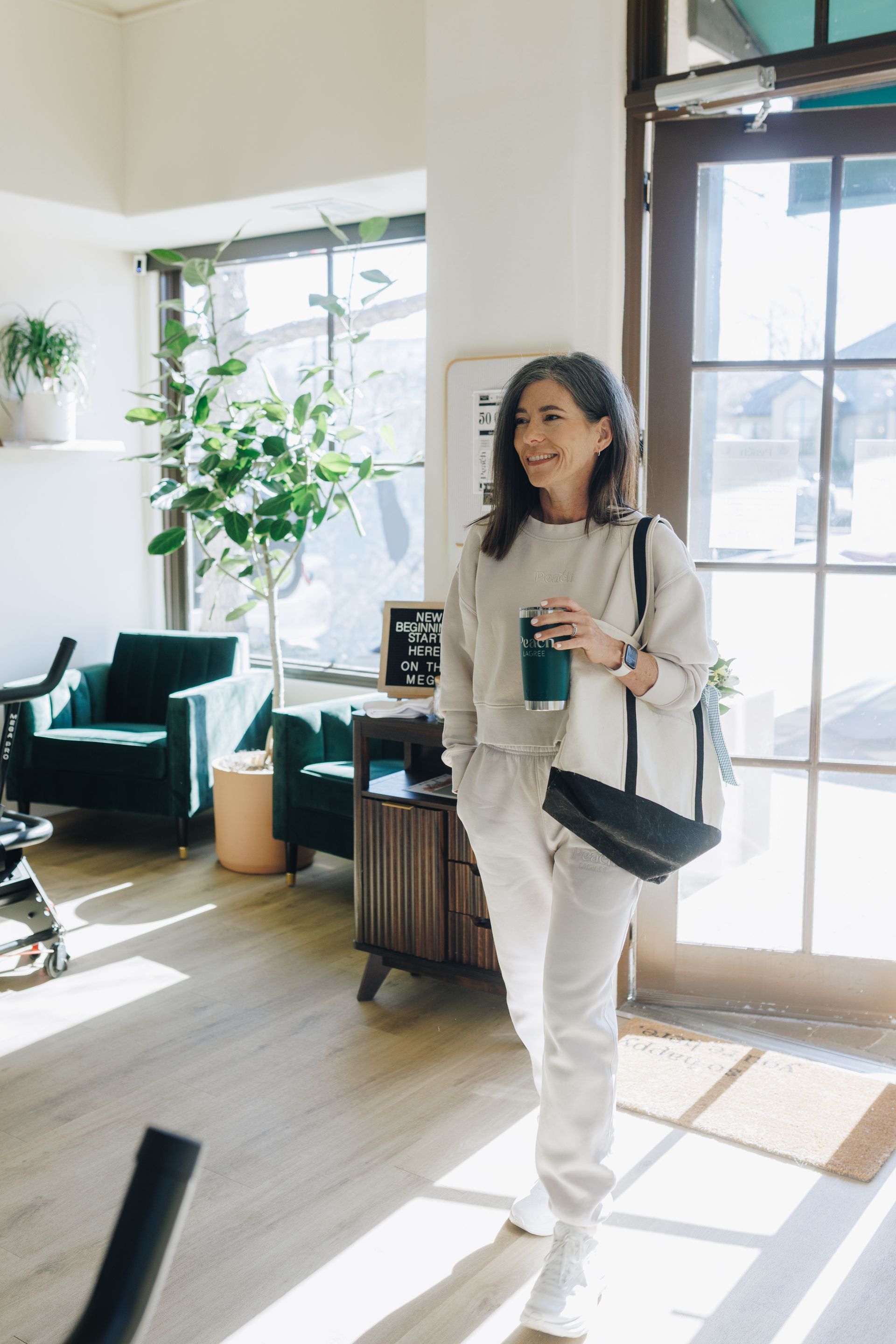 Woman in a bright lobby holding a drink and tote bag, standing near large windows and plants