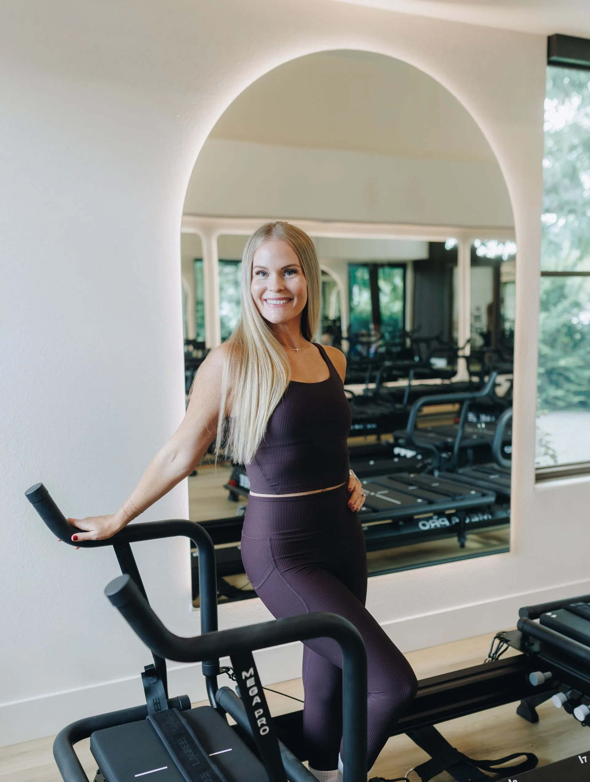 Woman posing on an exercise bike in a gym with large mirror and archway.
