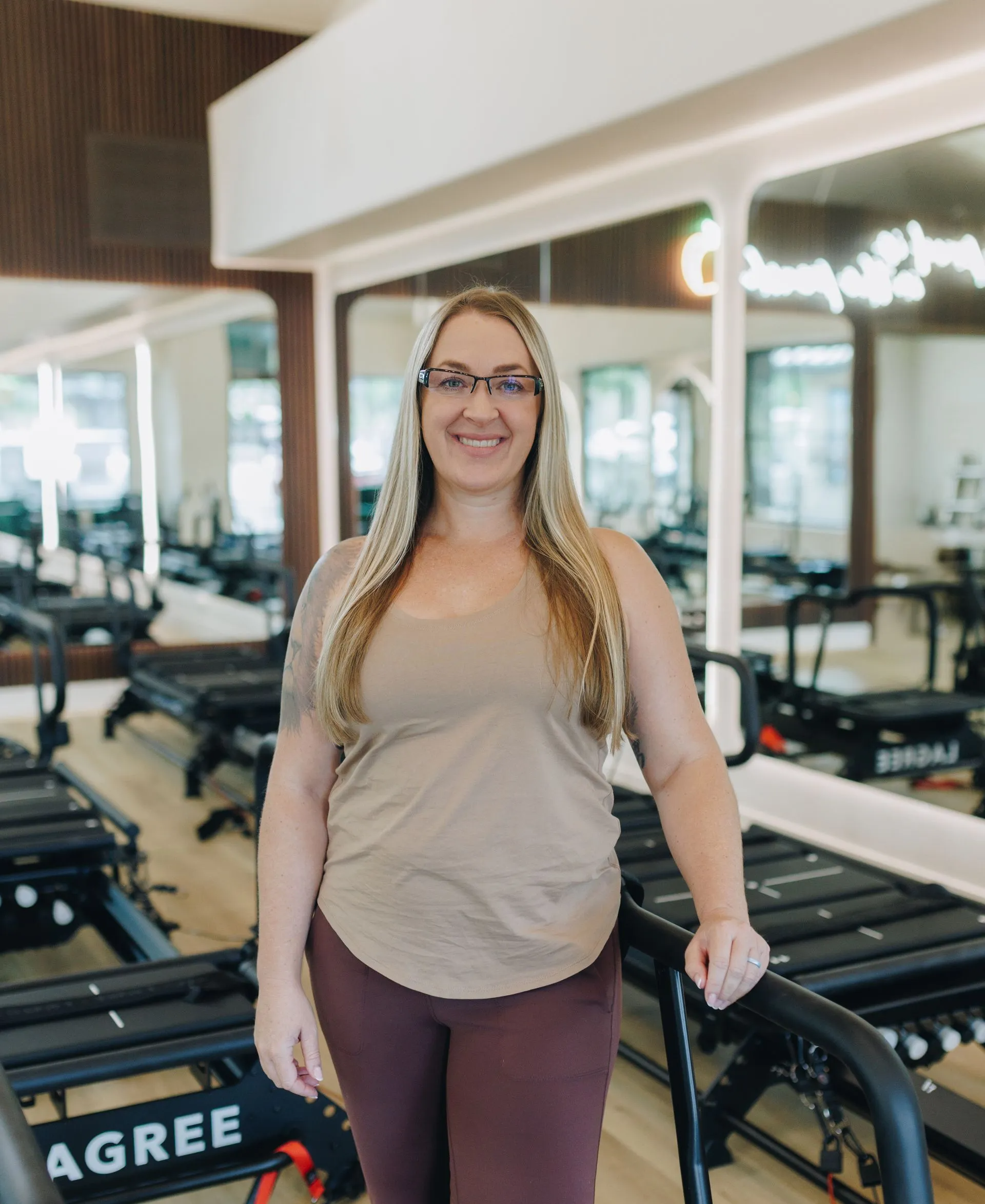 Woman standing in a gym beside rowing machines, smiling at the camera.