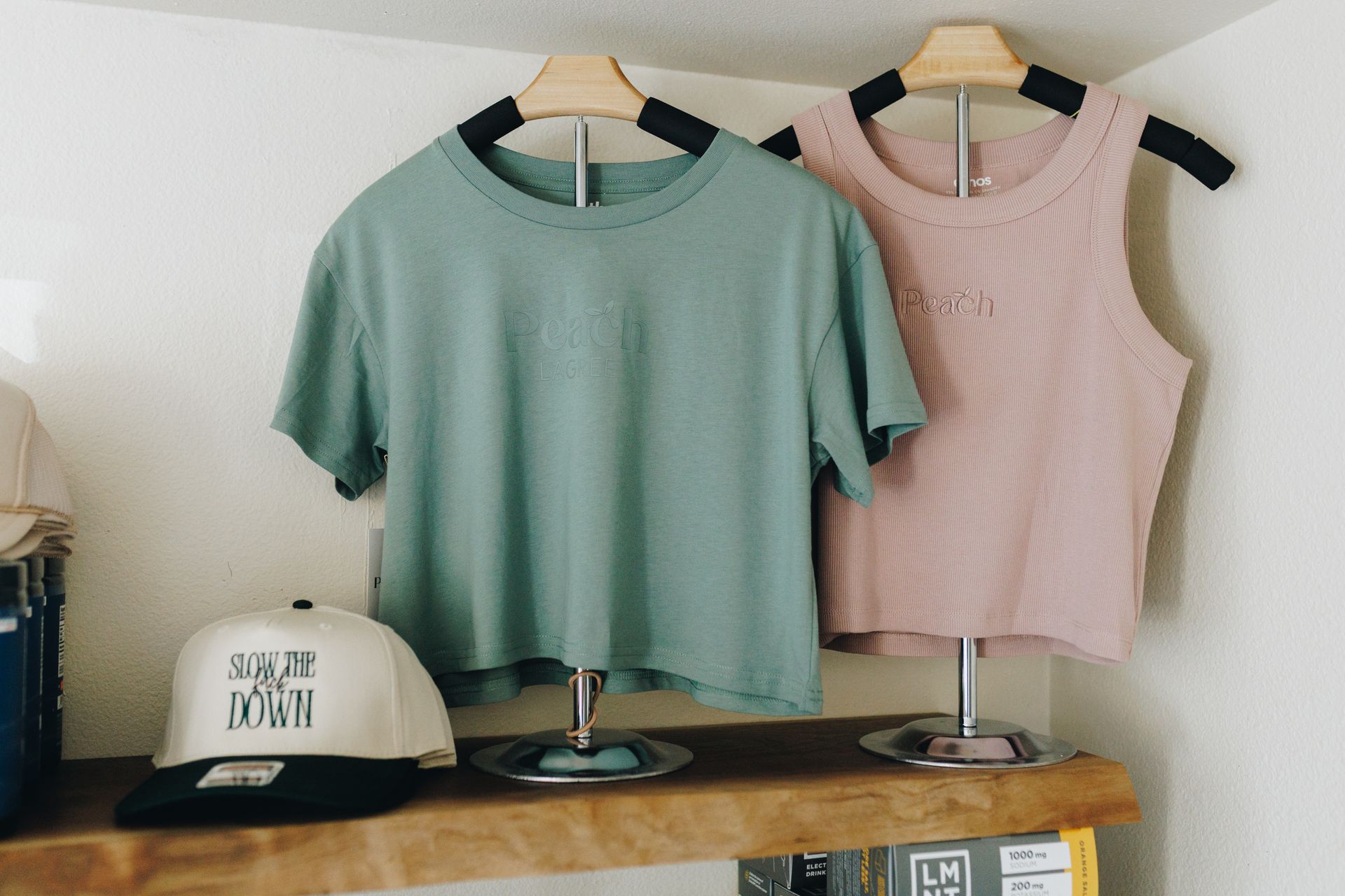 Two sleeveless tops on hangers, mint green and pink, displayed above a wooden shelf.