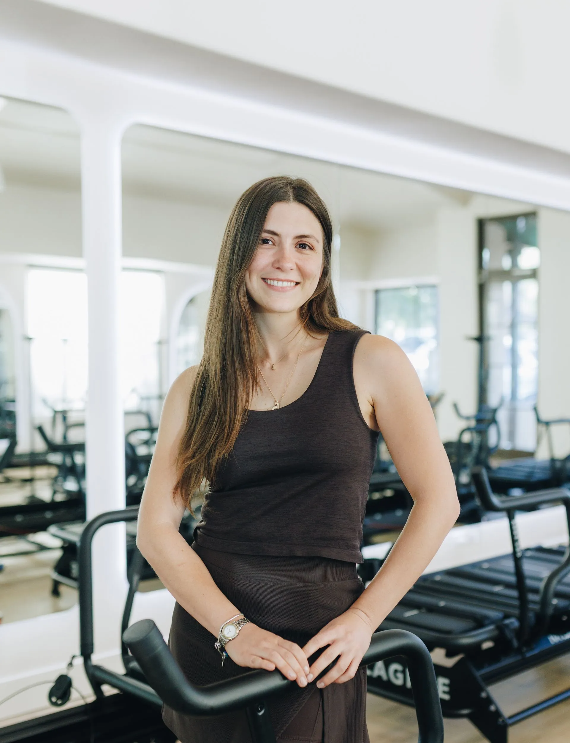 Woman smiling beside gym equipment in a bright fitness studio, wearing a dark tank top.
