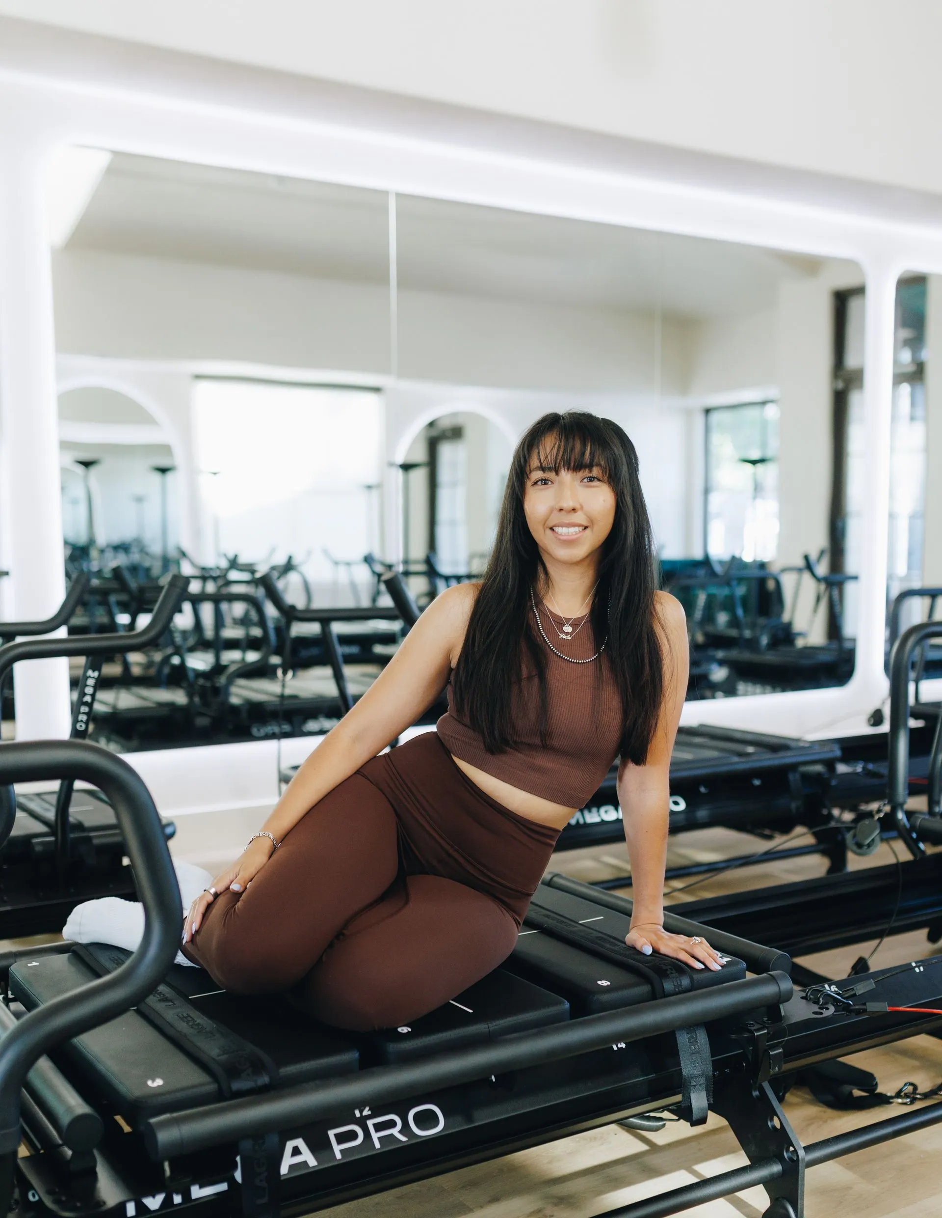 Woman posing on a reformer Pilates machine in a mirrored gym studio.