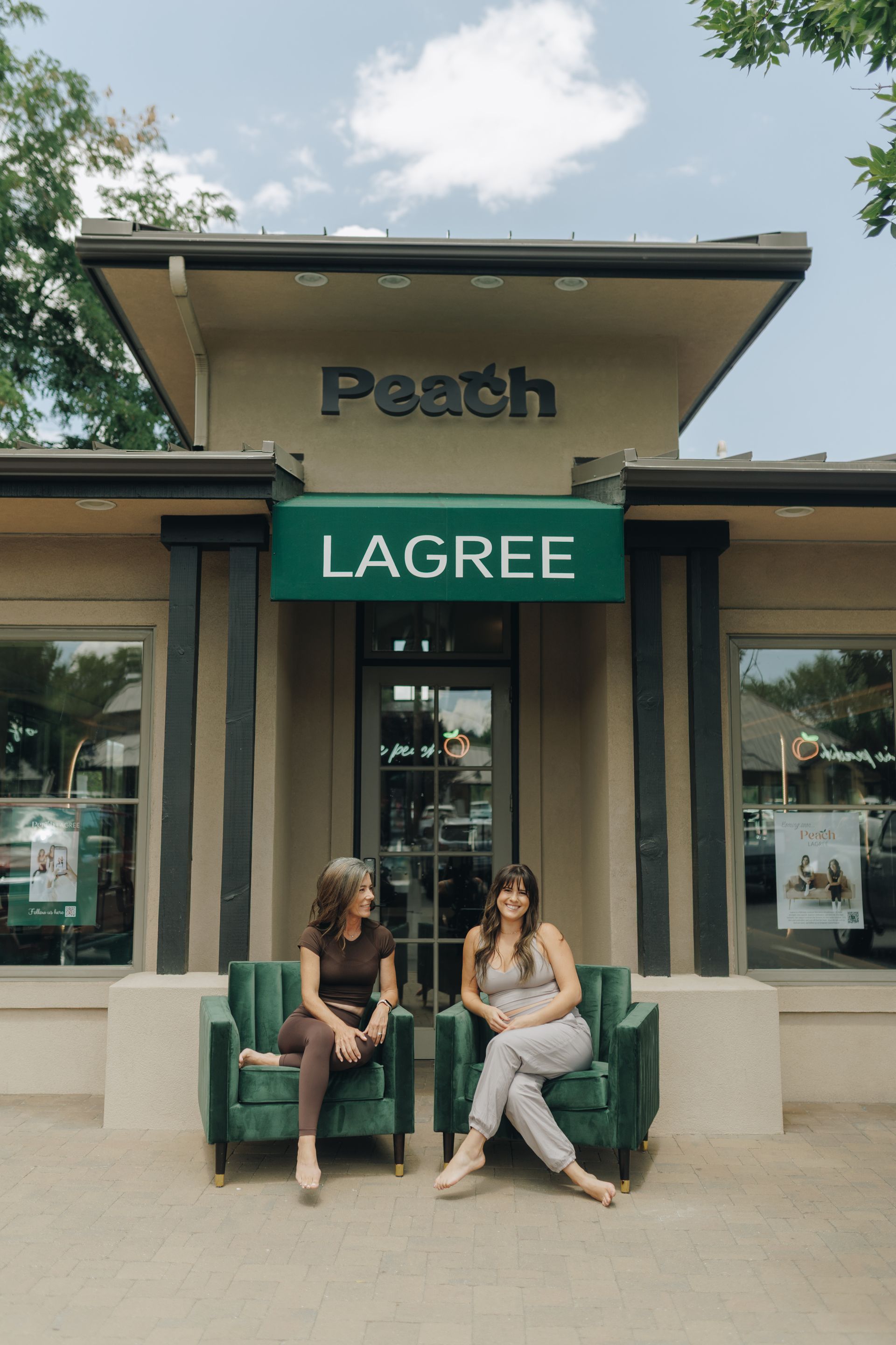Two people sitting outside a Lagree studio with a green sign and peach-colored storefront