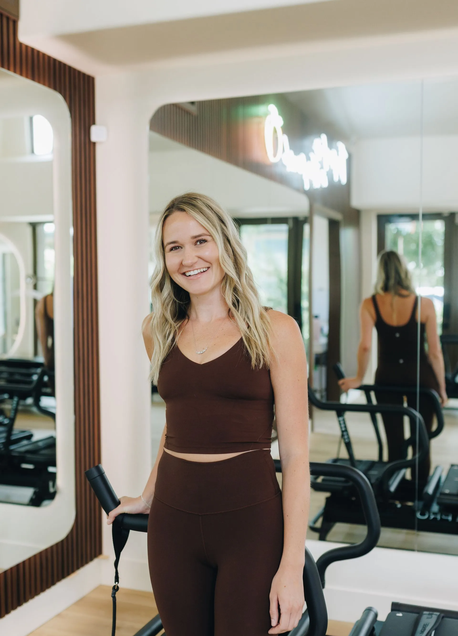 Woman in brown workout clothes standing in a gym, smiling near a mirrored wall and weight machines.